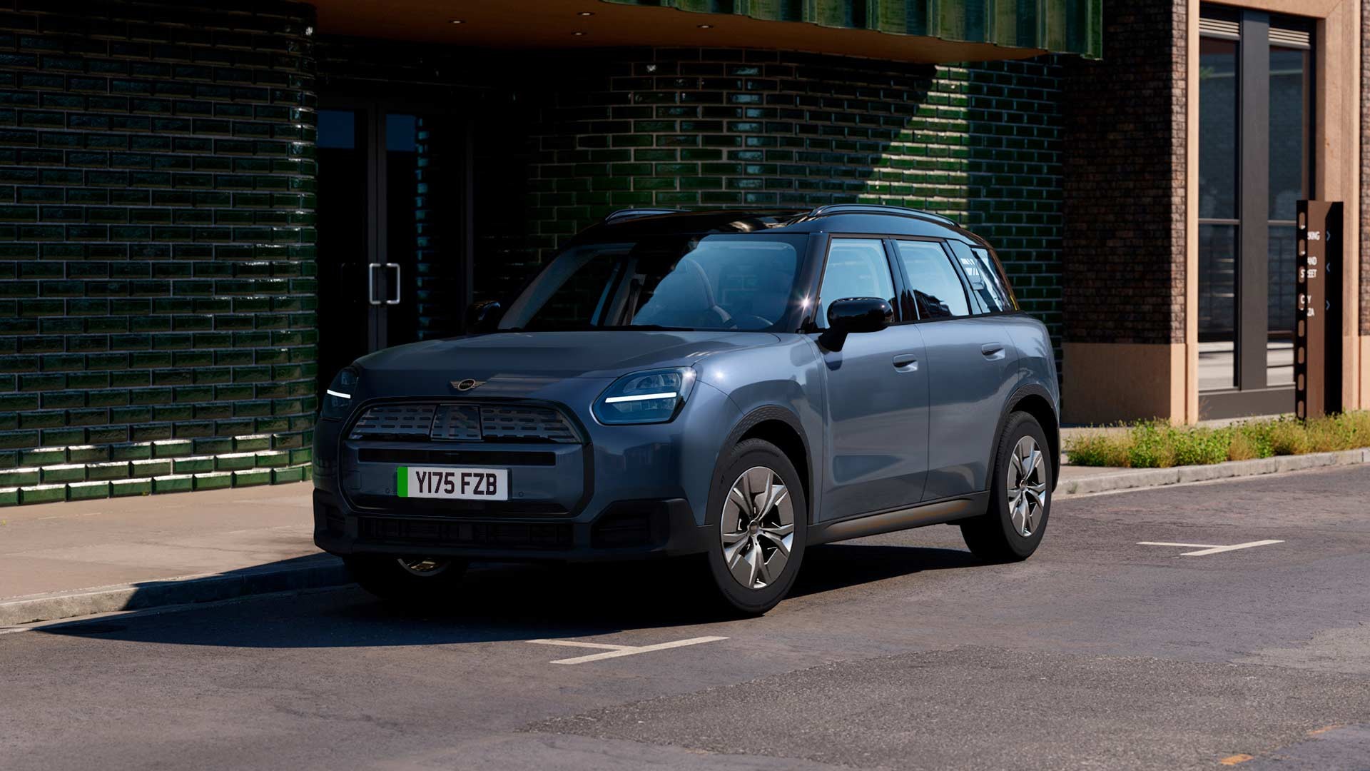 MINI Countryman electric SUV parked on a city street in front of a dark green tiled building, featuring black roof and modern headlights.